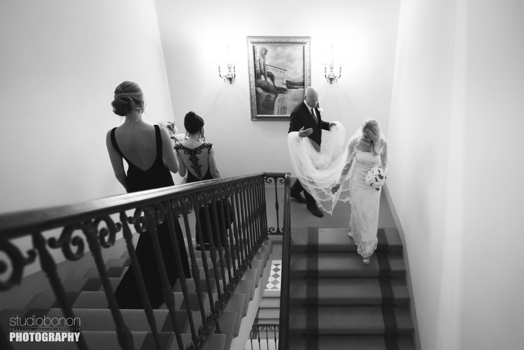 Bride Amy walks down hotel Four Seasons staircase with father and black dress elegant bridesmaids